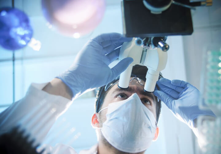 Scientist wearing a face mask and gloves examining samples through a microscope in a laboratory, with green-tinted lab equipment in the foreground.