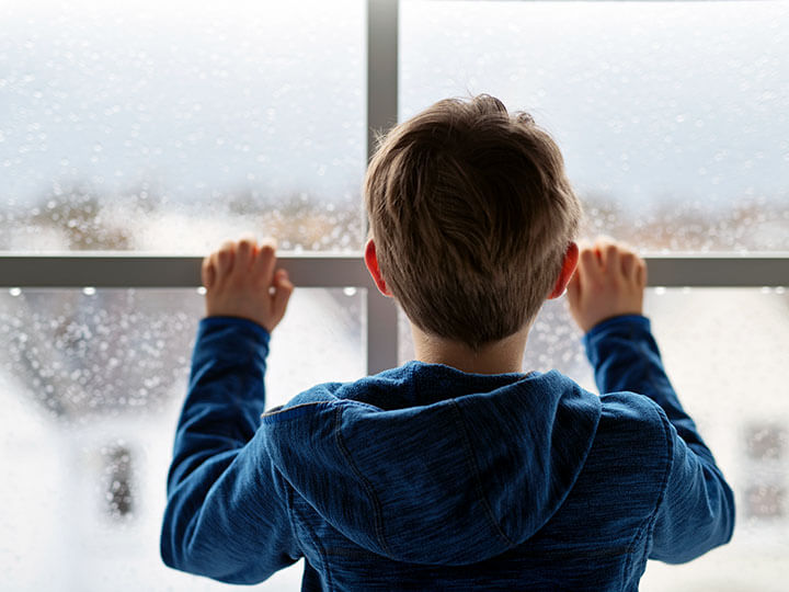 Rear view of young boy wearing a royal blue heather hooded sweatshirt looking out a window on a rainy day