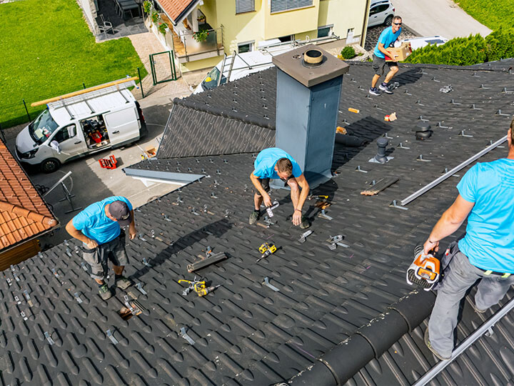 Aerial view of roofing crew of four males in blue shirts installing solar panels on a black shingle rooftop