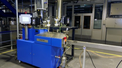 Industrial polymer compounding machine in blue and white, positioned within a factory environment filled with equipment.