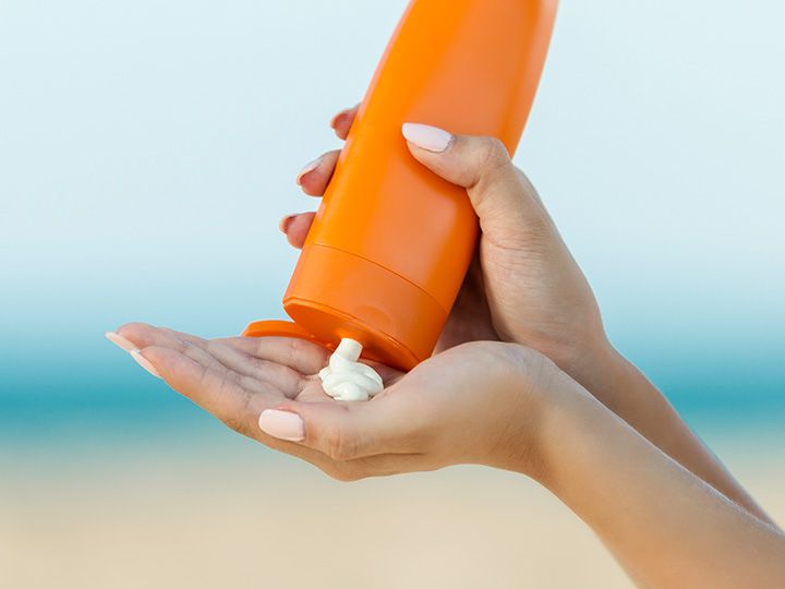A woman's hand hold an orange bottle of sunscreen cream, indicating a focus on sunscreen testing and safety.