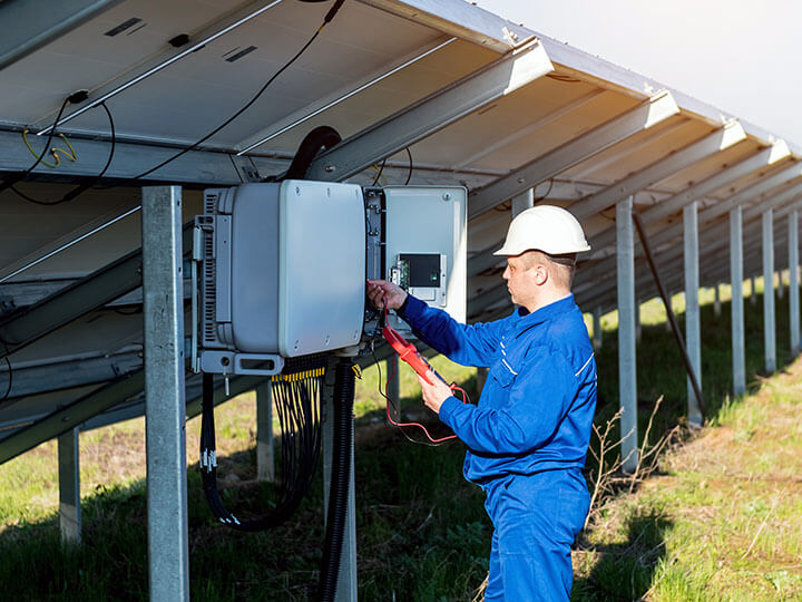 Male inspector in blue outfit and white hard hat checking the voltage output of an inverter for solar panels