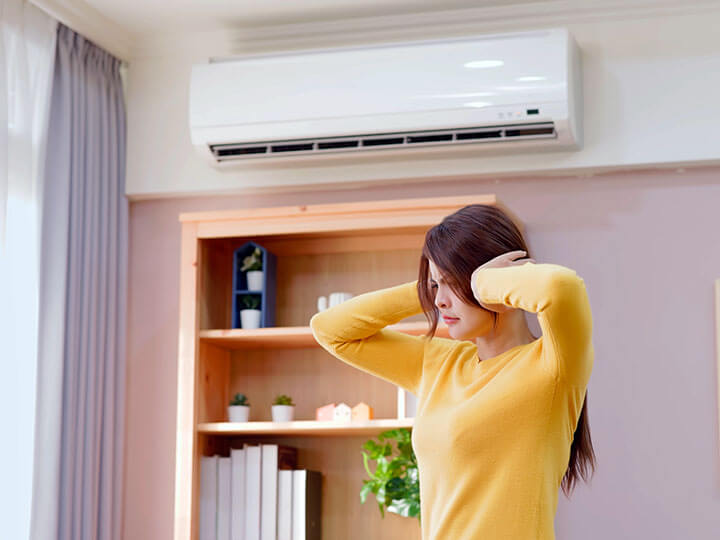 Asian woman in a yellow sweater covering her ears from the noise of the broken air conditioning unit on the wall behind her