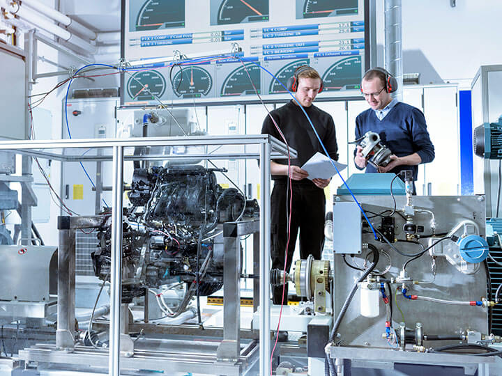 Two men examining an automotive component in an automotive research lab with engine hooked up to performance testing machine and monitors in background