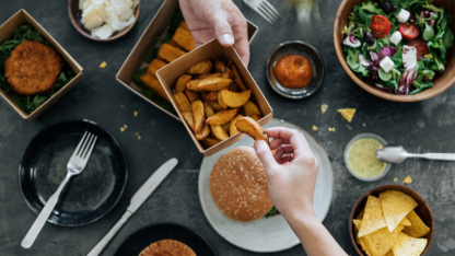 A person shares potato wedges from a box at a table set with a burger, salad, and other assorted foods.