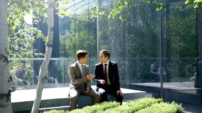 Two men in suits sit on a bench, facing a large glass wall, engaged in conversation.