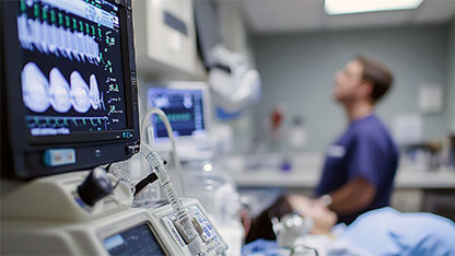 Close up view of Medical imaging and vitals machine and monitor with a medical staff member standing next to a patient in a bed in the background