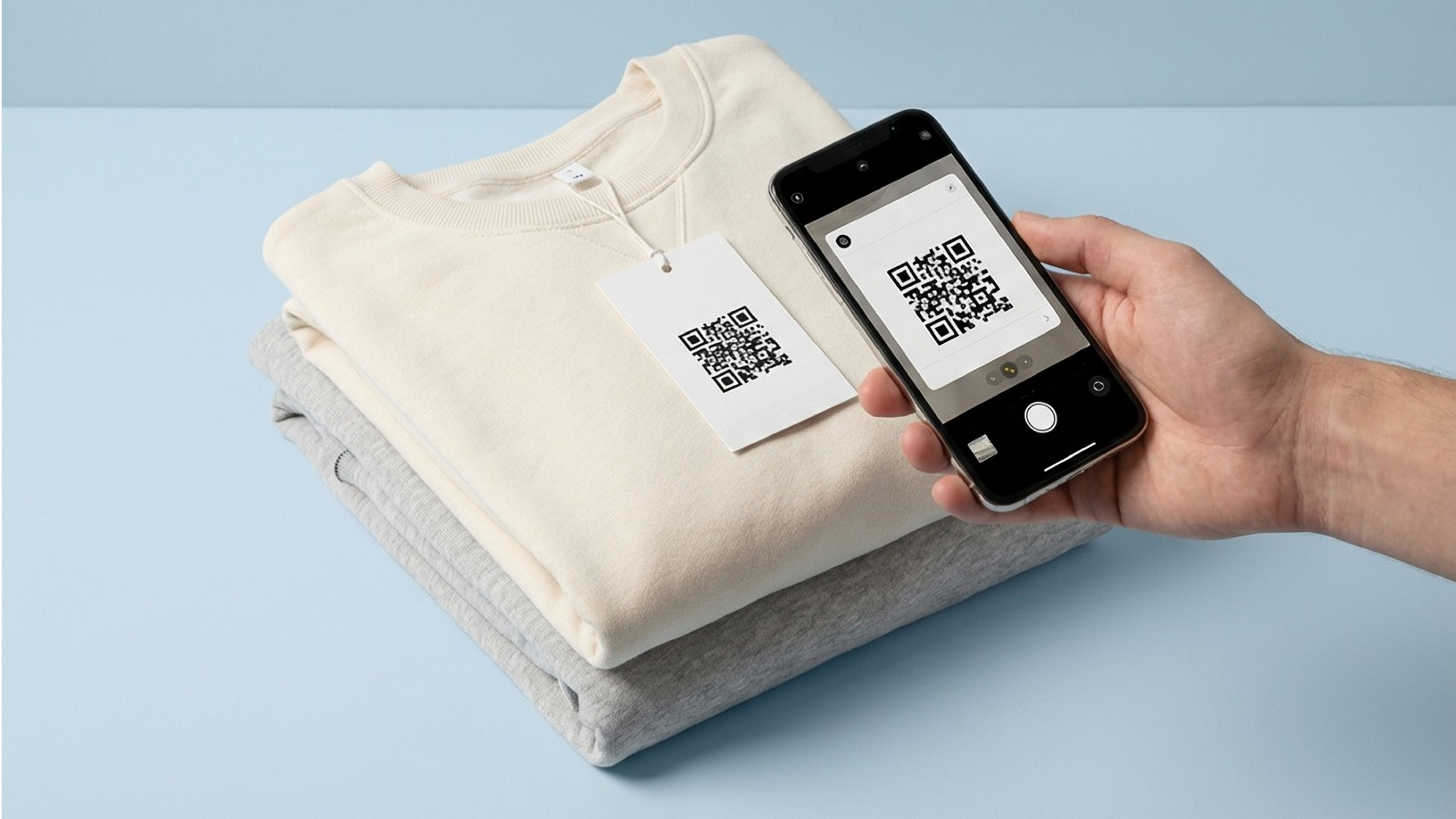 A comfy clothing displayed on a flat surface of a light blue color, a tag with QR code is attached to it. A phone is scanning the QR code on the tag.